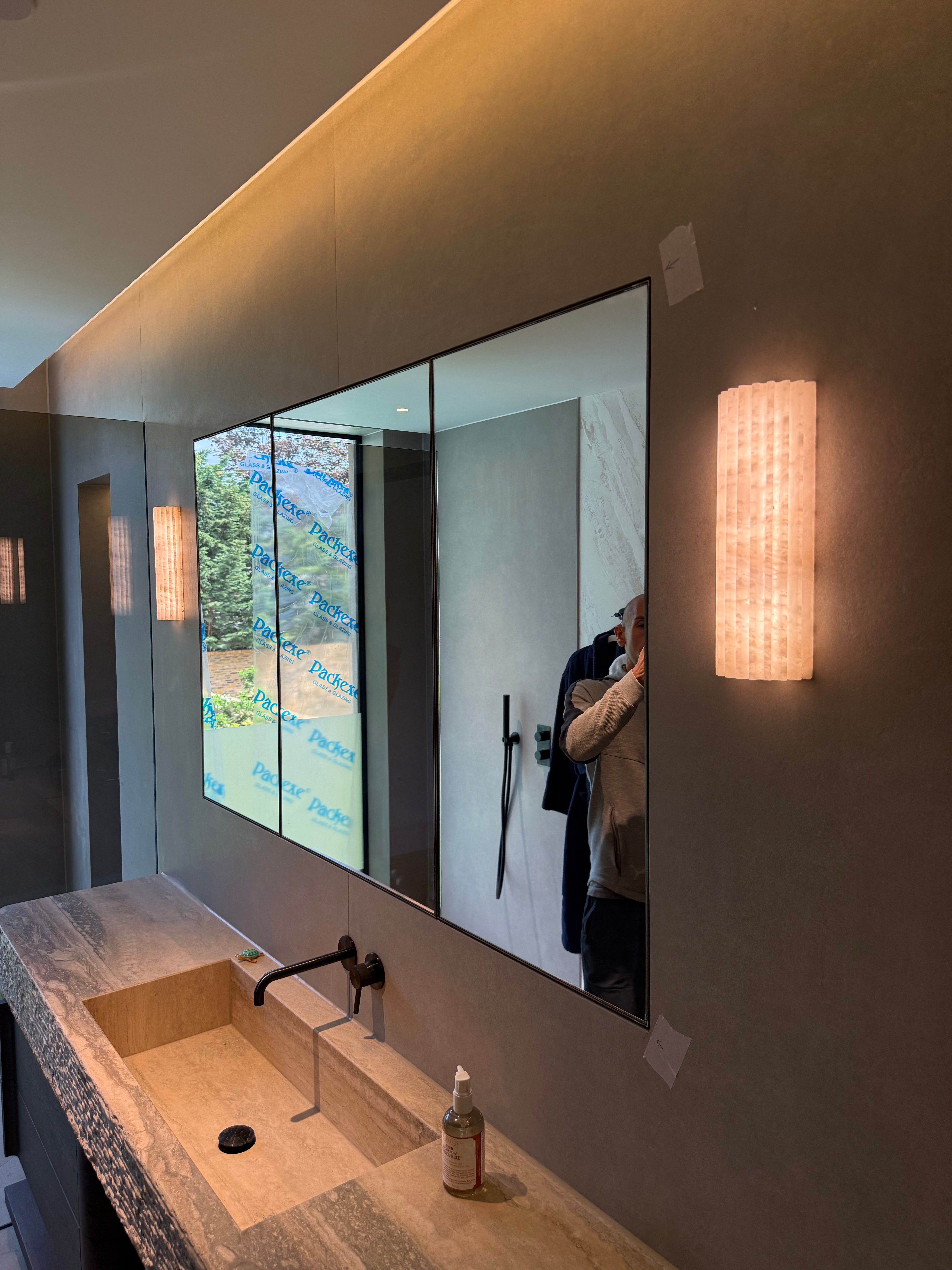 Bathroom with a large mirror reflecting a person taking a photo,  marble wall lights, and a sink.